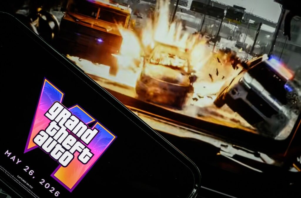 In this photo illustration, a phone displaying the Grant Theft Auto VI logo and new release date is shown in front of a screen displaying the second official trailer for the game.