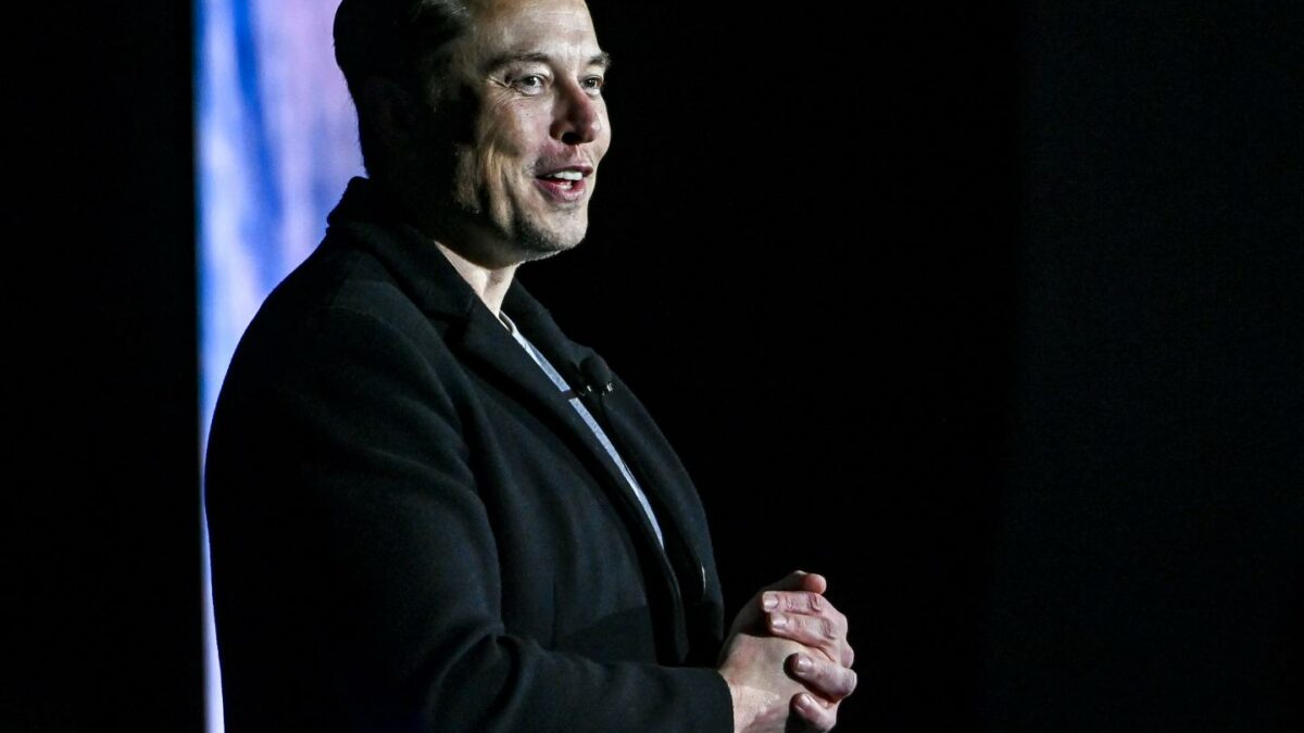 Elon Musk, wearing all black, smiles and looks to the side.