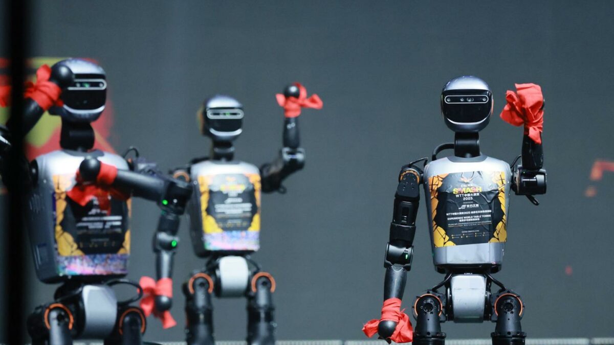 A group of three robots waiving hello to the audience from a stage.
