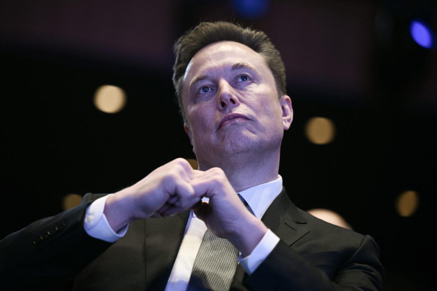 Elon Musk sits with his fists together, looking up.