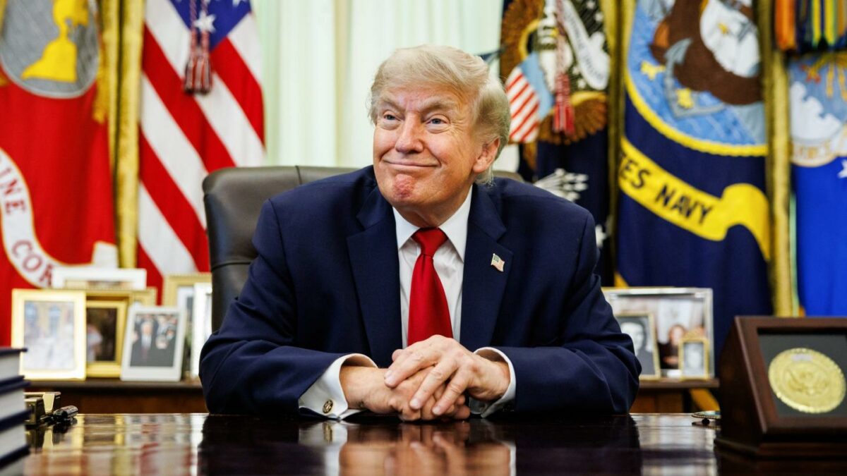 Donald Trump sits at his desk in the Oval Office, smiling and with his hands folded in front of him.