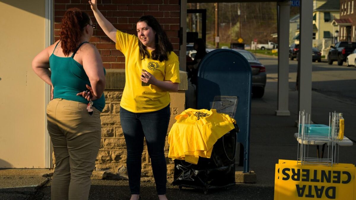 A woman in a yellow jacket in front of a "NO DATA CENTERS" sign, speaks with another woman with her hands on her hips.