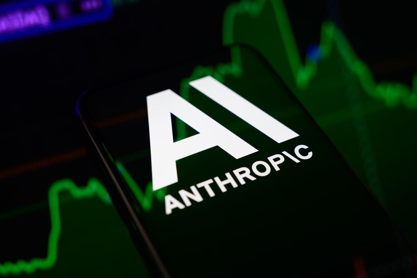 The Anthropic logo is displayed on a smartphone screen placed on a reflective surface onto which a stock market chart is projected.